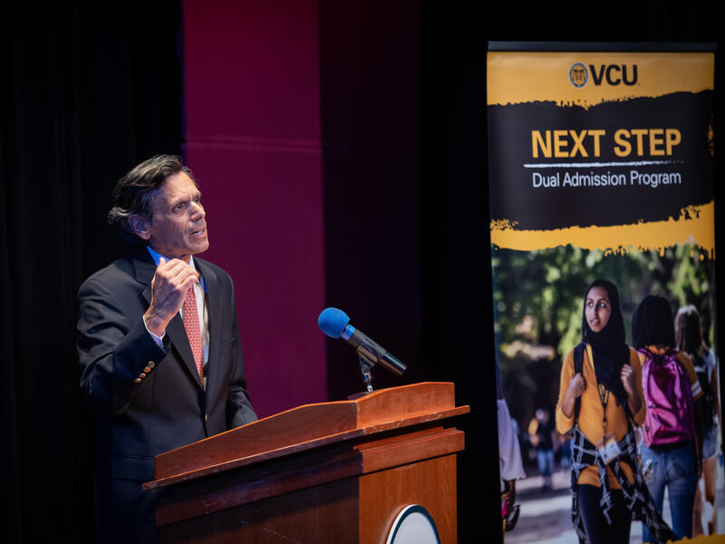 A man in a coat and tie gestures while speaking at a lectern into a microphone.