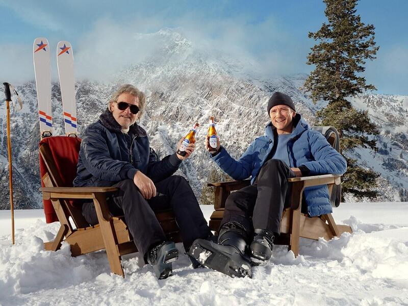 A photo of two men sitting on a snowy moutain in wooden chairs. Each man is holding a Michelob ULTRA beer. Behind the man on the left is a pair of skis. 