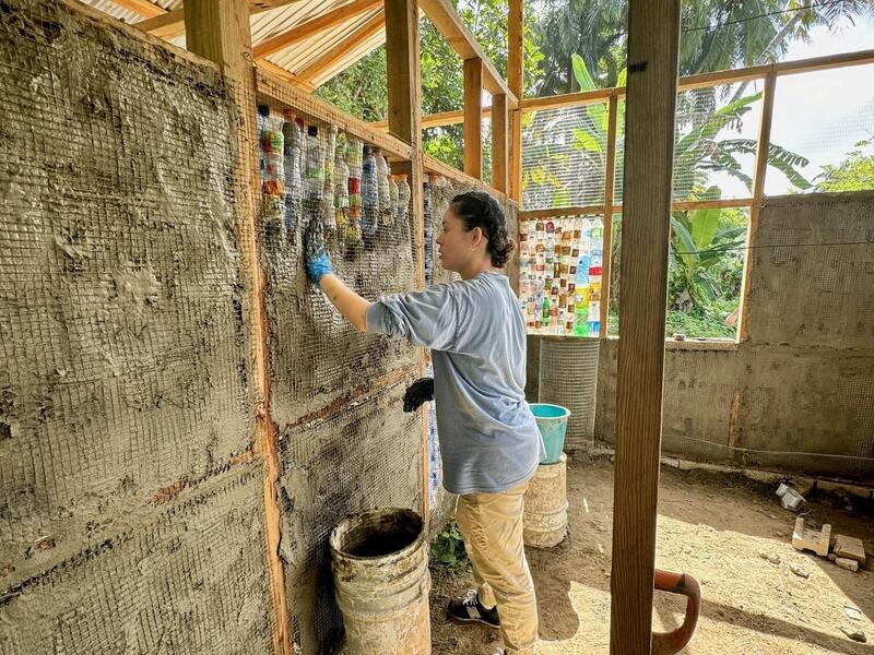 A photo of a person spreading mud over water bottles and chicken wires in between wooden frames for a house