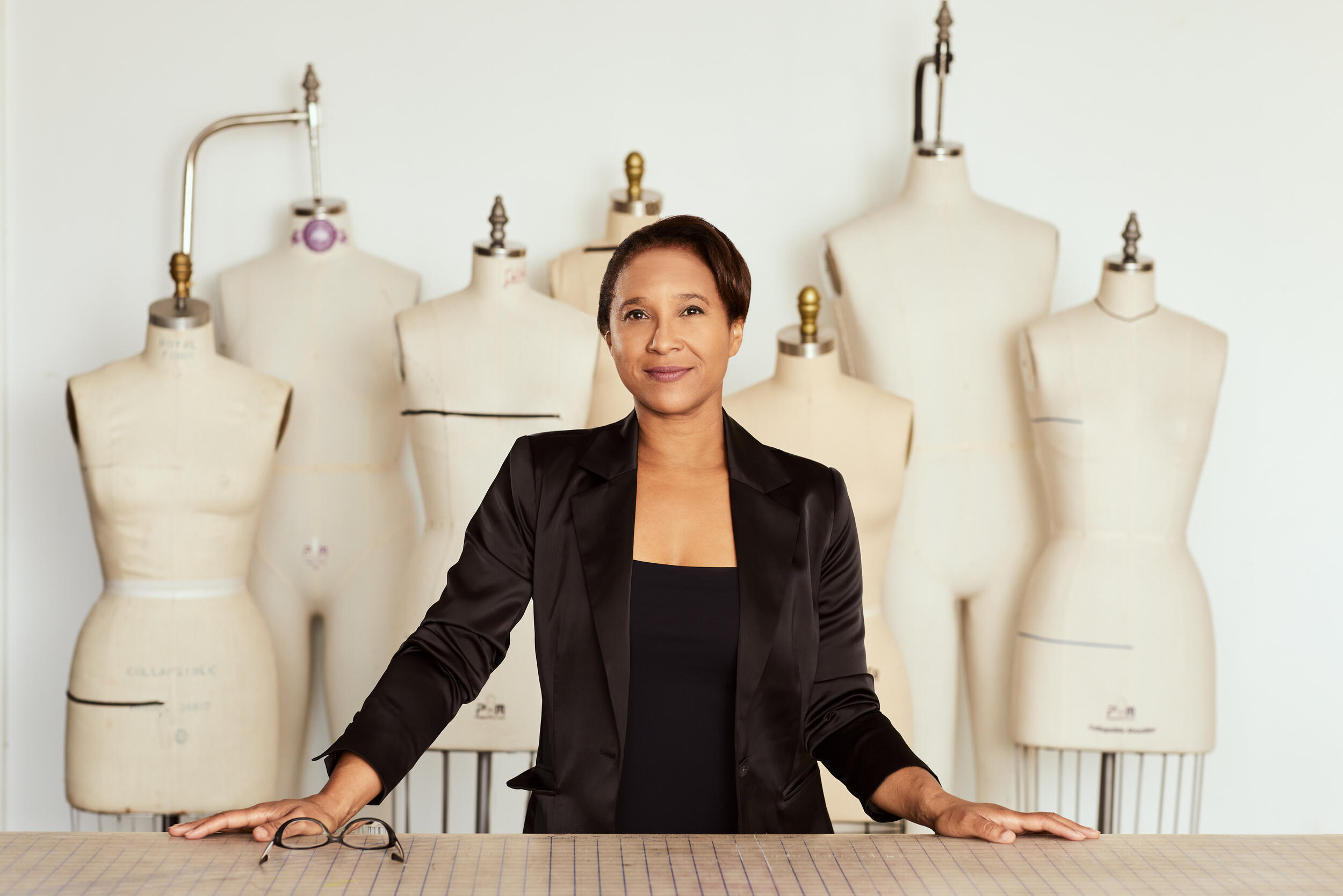 A photo of a woman from the waist up standing behind a table. Behind the woman are seven sewing mannequins.