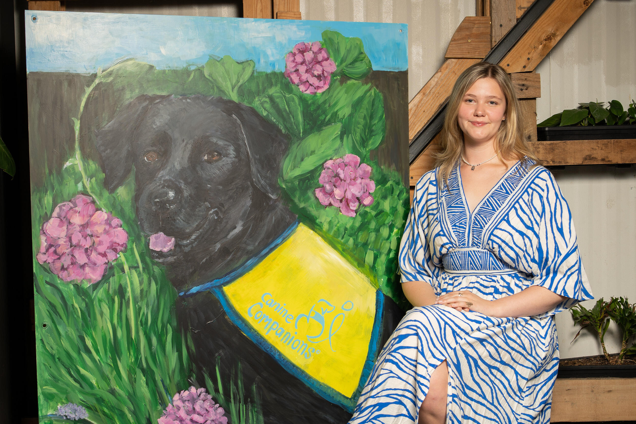 A photo of a woman sitting to the right of a painting of a dog. 