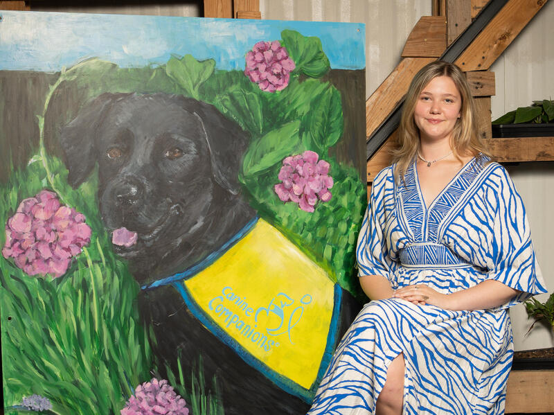 A photo of a woman sitting to the right of a painting of a dog. 