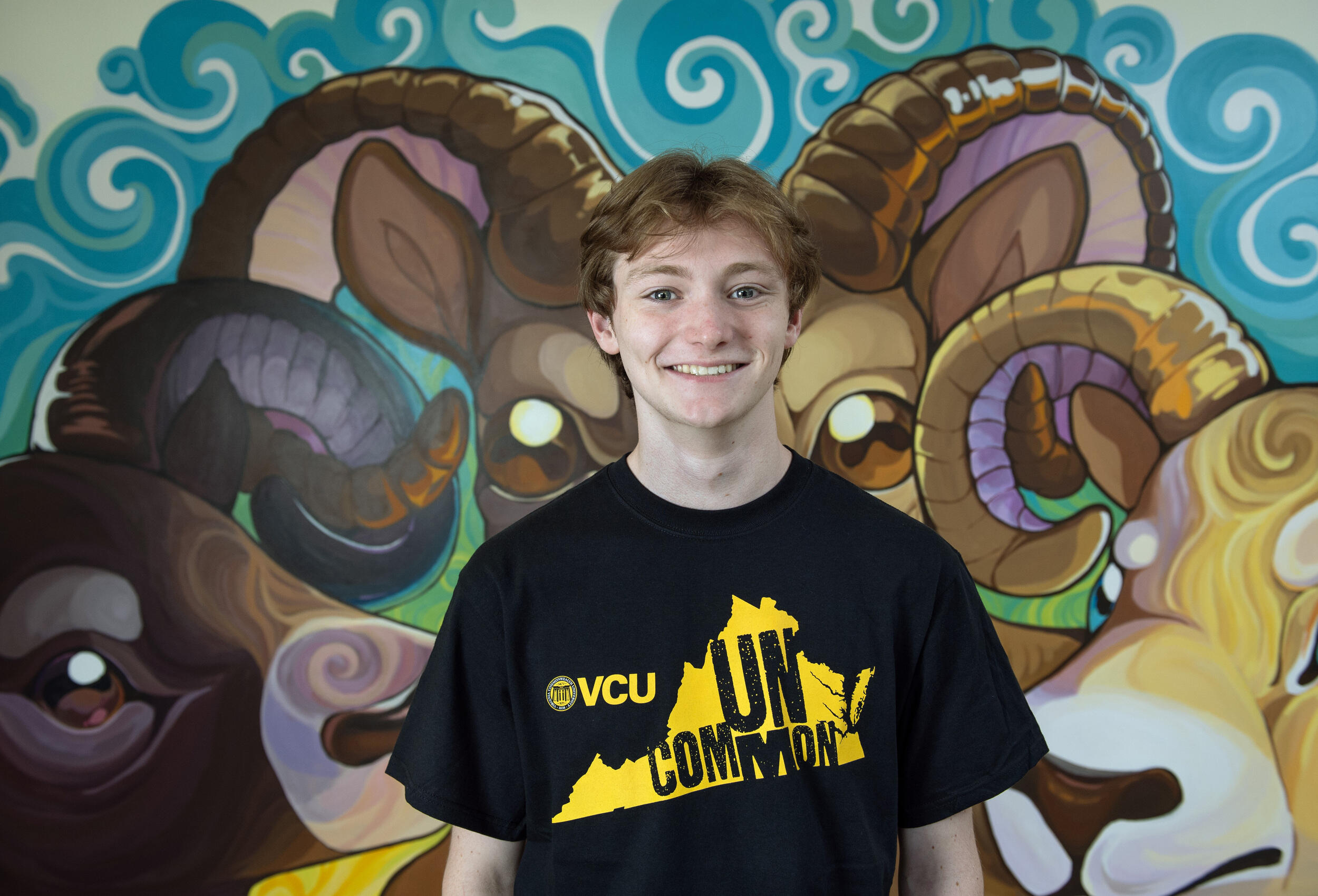 A photo of a man wearing a black t-shirt from the chest up. His t-shirt has the shape of Virginia printed on it. Inside the state's silhouette is black text that reads \"UNCOMMON.\" Behind the man is a mural of three rams. 