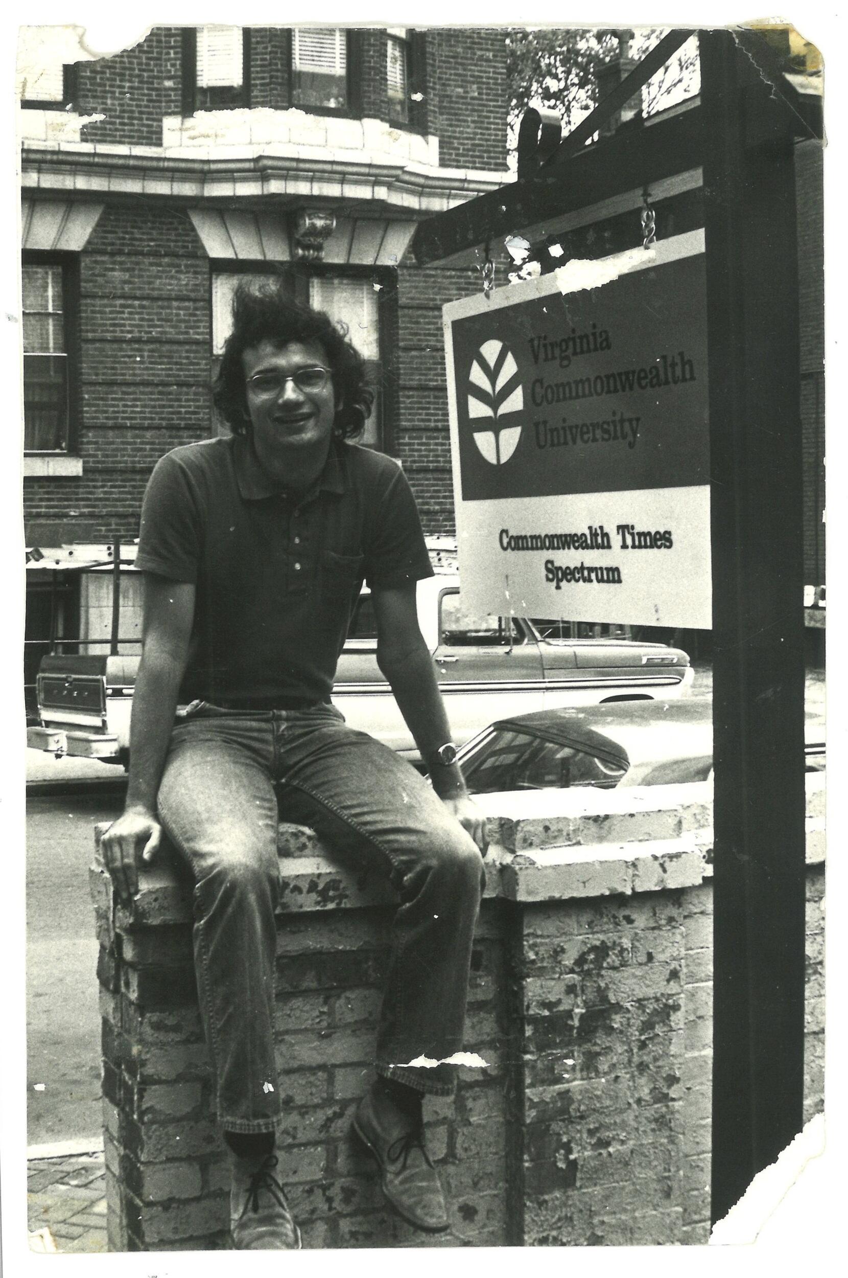 A black and white photo of a man sitting on a brick fence. Next to him is a sign that says \"Virginia Commonwealth University, Commonwealth Times, Spectrum.\" 