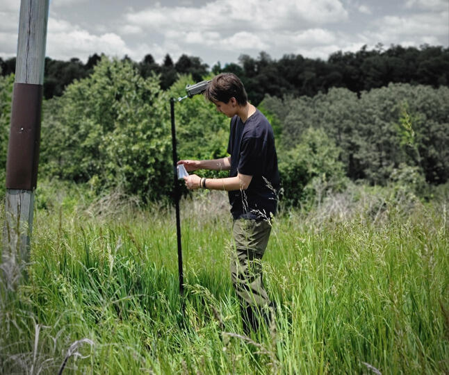 A photo of a person setting up a trail camera in a field of tall grass next to a treeline.
