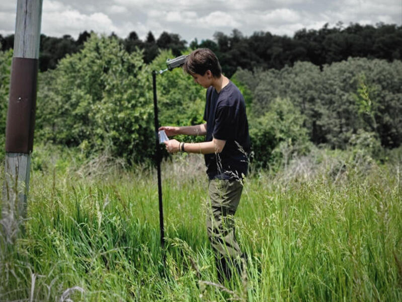 A photo of a person setting up a trail camera in a field of tall grass next to a treeline.