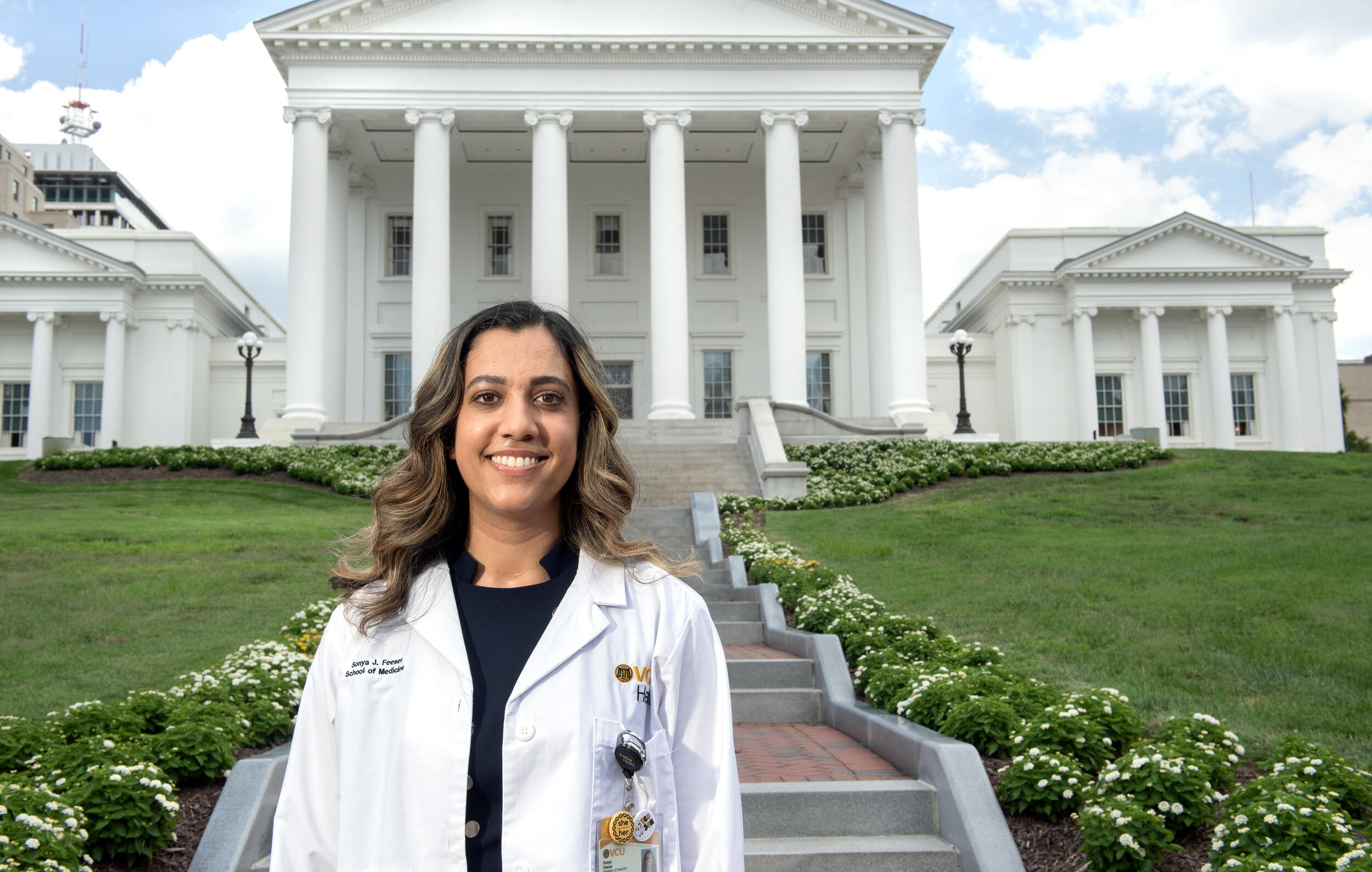 A photo of a woman wearing a white doctor's coat from the chest up. She is standing in front of a stair case outside. The staircase leads to the Virginia State Capitol. 