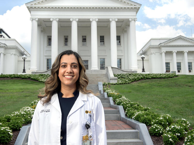 A photo of a woman wearing a white doctor's coat from the chest up. She is standing in front of a stair case outside. The staircase leads to the Virginia State Capitol. 