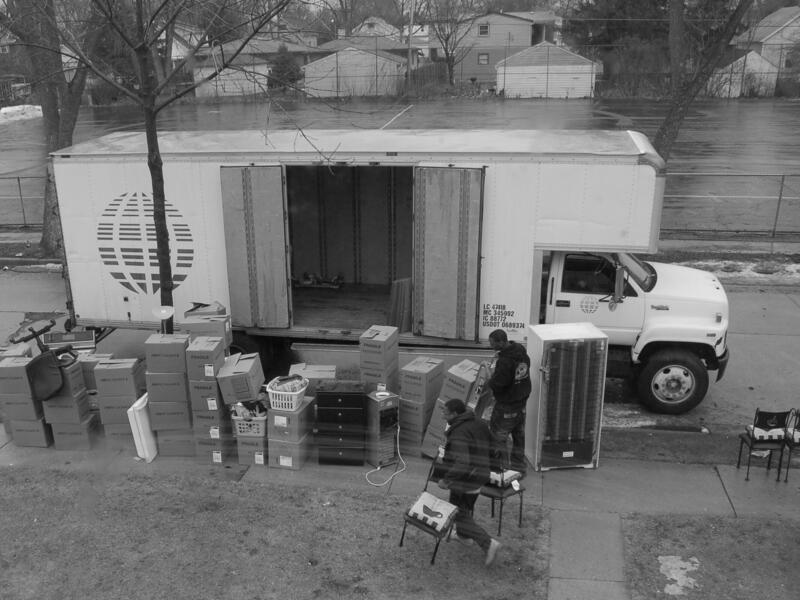 A moving van with its side door open is parked on a street. Various household items and moving boxes sit alongside it. Two moving assistants are putting items next to the van.