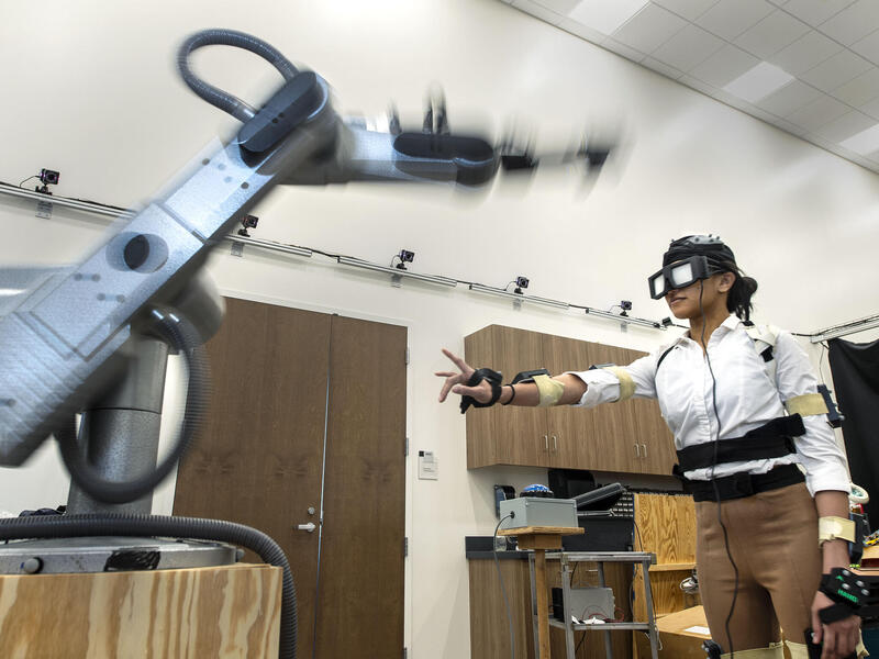 Person in motion-capture suit reaches out her hand toward a robotic arm.