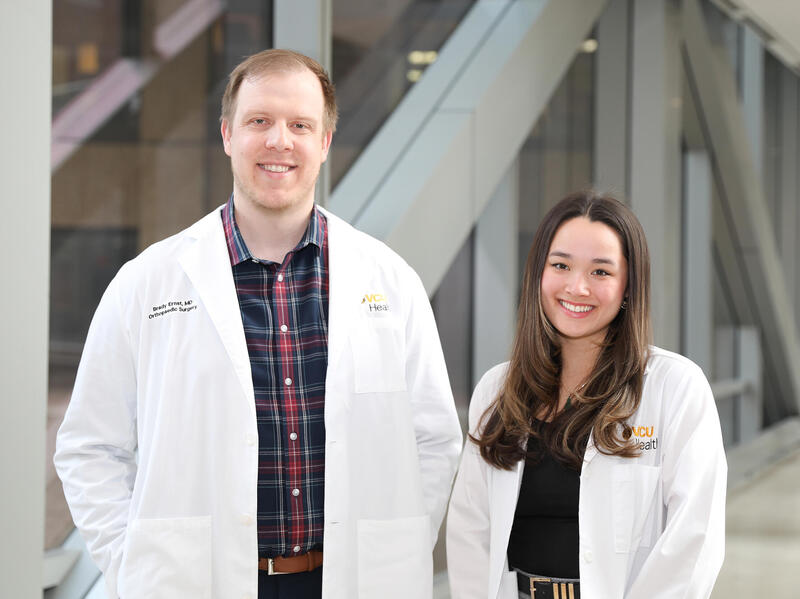 A photo of a man and a woman standing. Both are wearing white medical coats. 