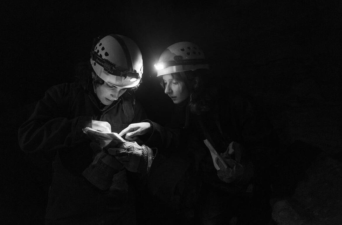 A photo of two people wearing helmets in the dark looking at a map. The two are only illuminated by the lights on their helmets. Because of this, you can only see their faces, hands, and a bit of their torso. 