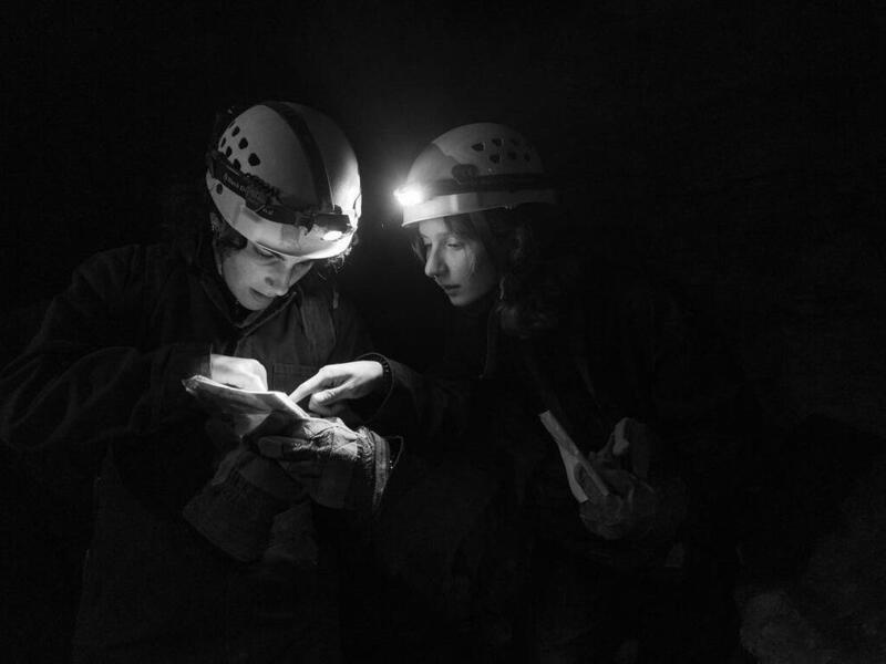 A photo of two people wearing helmets in the dark looking at a map. The two are only illuminated by the lights on their helmets. Because of this, you can only see their faces, hands, and a bit of their torso. 