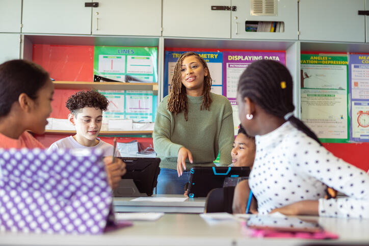 A photo of a teacher talking to four students sitting at two tables in front of her. The two boys are holding tablets. 