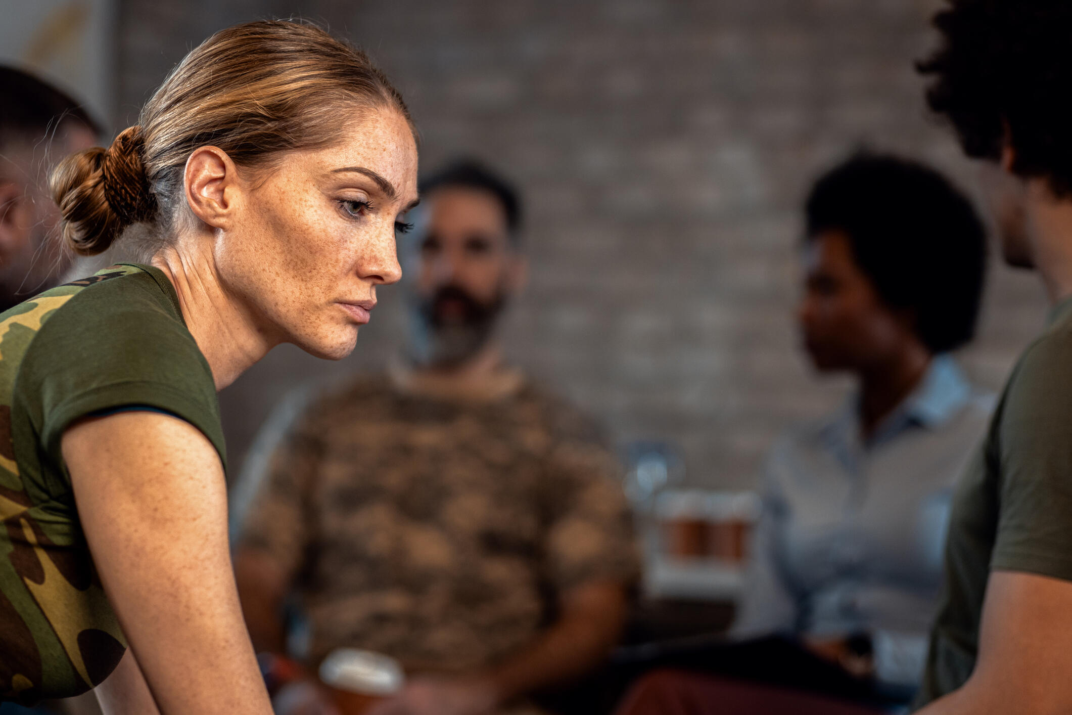 A photo of a woman looking to the right somberly. Behind her four people sit in a semi-circle talking to each other