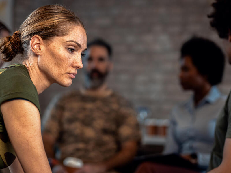 A photo of a woman looking to the right somberly. Behind her four people sit in a semi-circle talking to each other